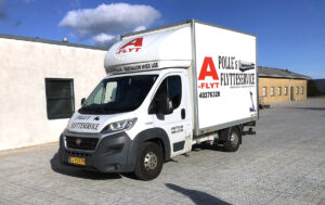 En hvid flyttebil med logoet "A-FLYT" parkeret på en grusbelagt gårdsplads. Blå himmel og bygninger i baggrunden.